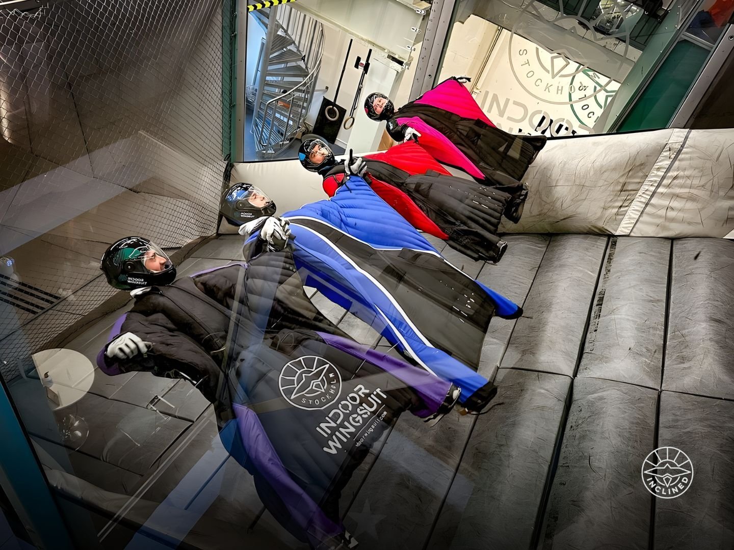 Multiple people in colorful wingsuits lying on a padded surface inside an indoor flight area.
