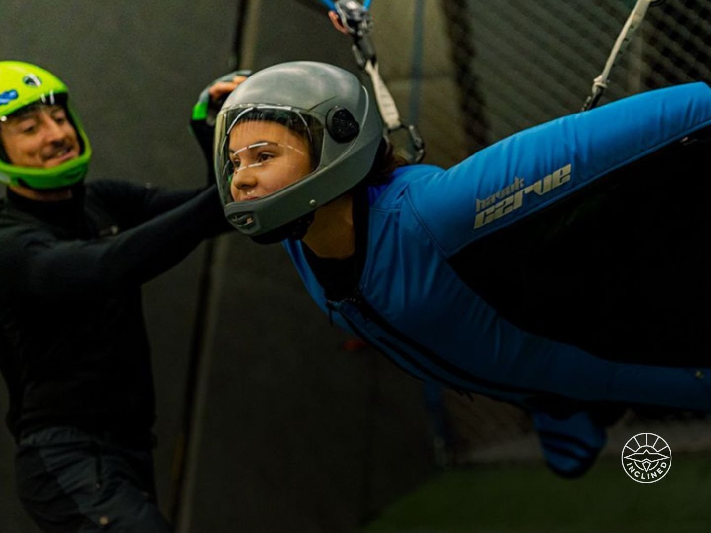 Child in a blue wingsuit flying indoors with an instructor guiding in a wind tunnel.