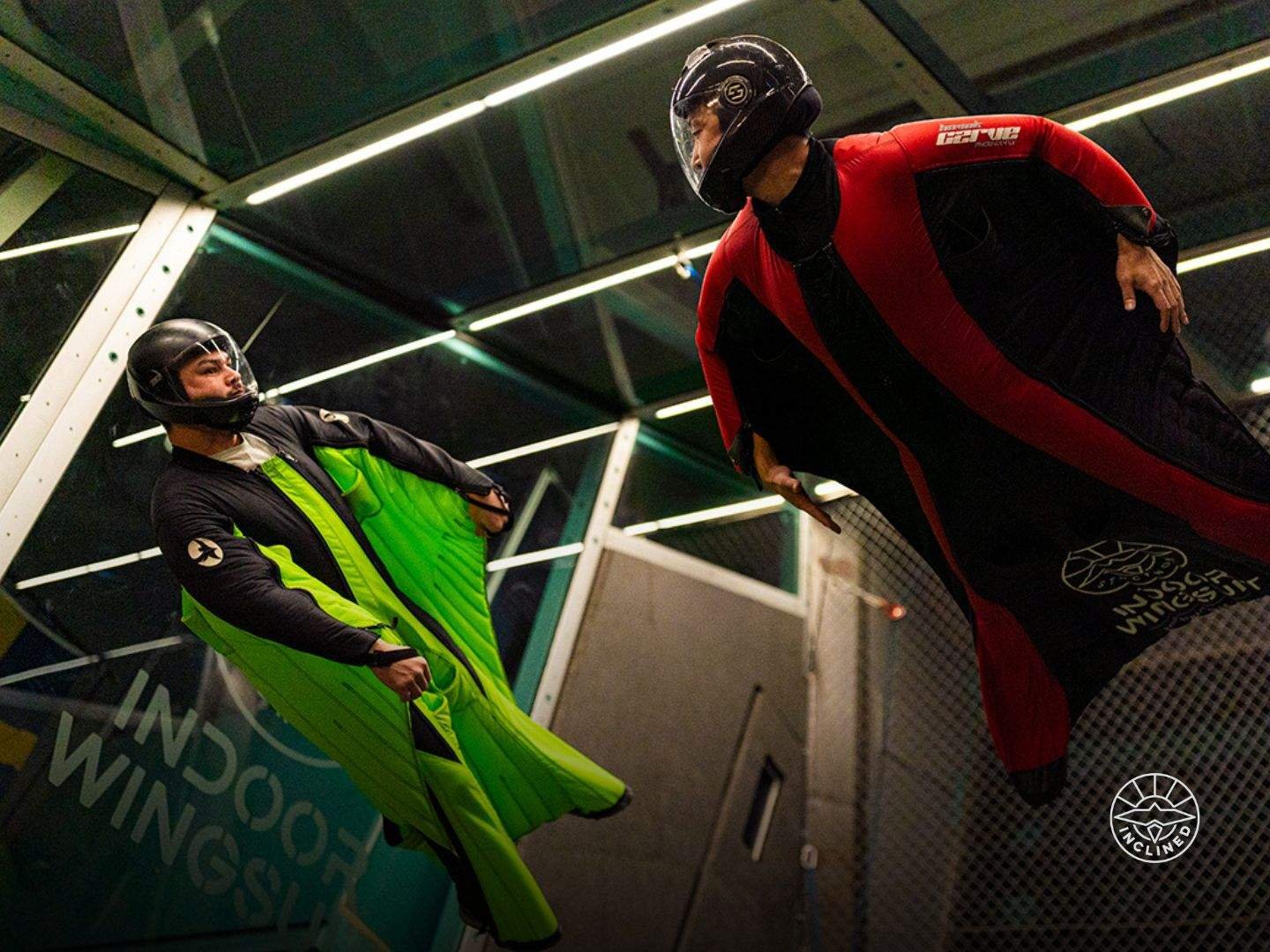 Two individuals in bright wingsuits practicing advanced maneuvers in a wind tunnel.